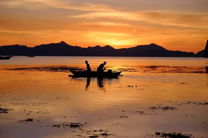 Dois pescadores num barco pequeno ao pôr do sol, com montanhas ao fundo