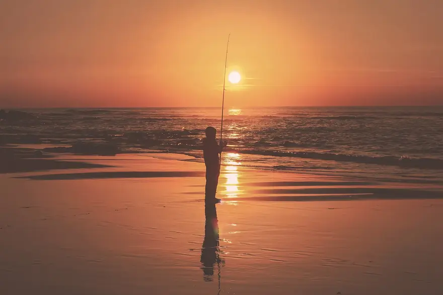 Pescador com cana comprida na praia ao pôr do sol